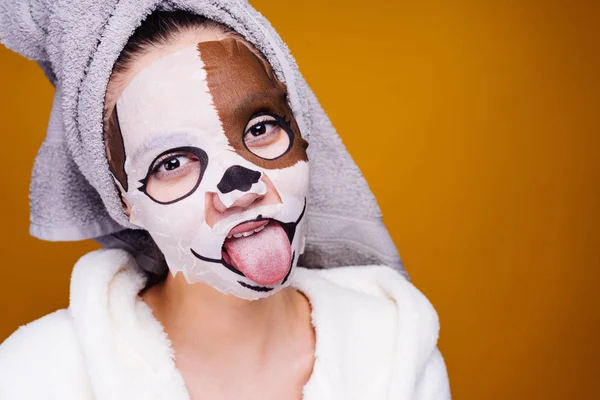 Een grappig meisje met een handdoek op haar hoofd draagt zorg voor zichzelf, toont haar tong, op haar gezicht een hydraterende masker met een snuit-hond — Stockfoto