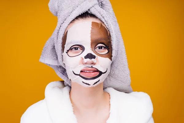 A young girl with a towel on her head and in a bathrobe after a shower looks after her face, a moisturizing mask with a snout dog — Stockfoto