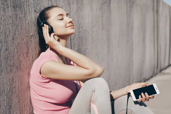 Tranquilla ragazza seduta a terra al sole, che si rilassa dopo un allenamento e ascolta musica sulle cuffie — Foto Stock