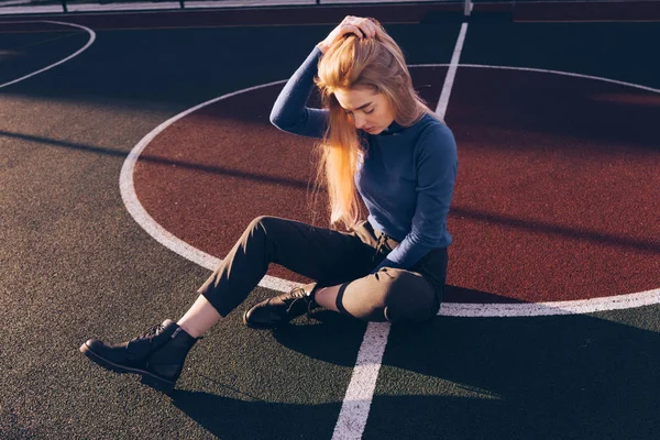 Elegante jovem loira modelo menina em camisola azul senta-se no campo de esportes no sol da manhã — Fotografia de Stock