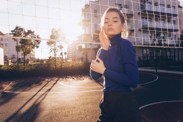 Modelo de menina bonita confiante em um suéter elegante posando em um campo de esportes nos raios do sol da manhã — Fotografia de Stock