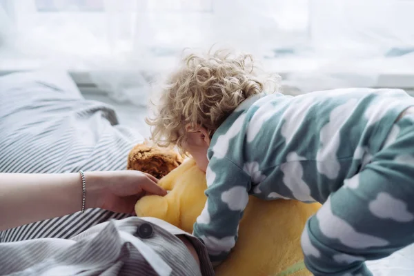 Кучерявий маленький хлопчик у блакитній піжамі грає з м'якою іграшкою поруч з мамою — стокове фото