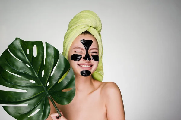 Una giovane ragazza ridente si prende cura di se stessa, sul suo viso una maschera nera contro l'acne, tiene una foglia verde — Foto Stock