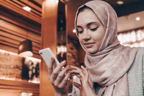 beautiful Arab girl in hijab at a cozy restaurant, writes a message on her smartphone