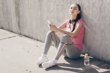 spor giyim sakin genç kız açık havada, yerde oturuyor dinlenme ve kulaklıklar müzik dinlemek