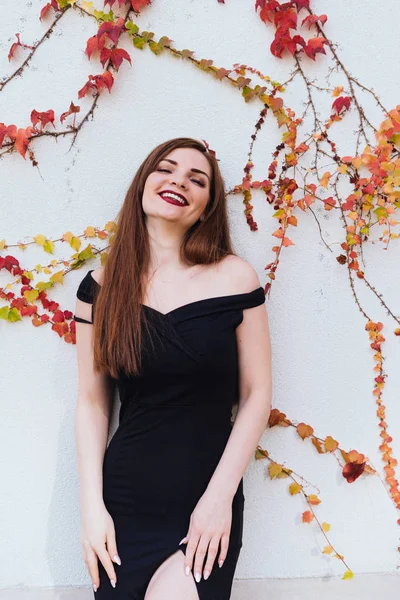 Atractiva chica de pelo largo en un vestido largo negro posando y riendo en la pared, disfrutando de una vida rica — Foto de Stock