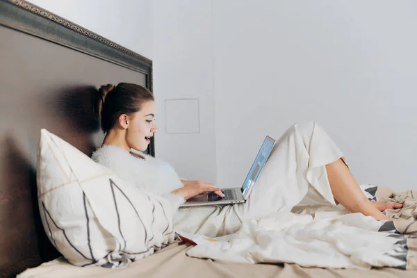 Joven mujer en pijama blanco se encuentra en la cama en su día libre, viendo la película en el ordenador portátil y riendo —  Fotos de Stock