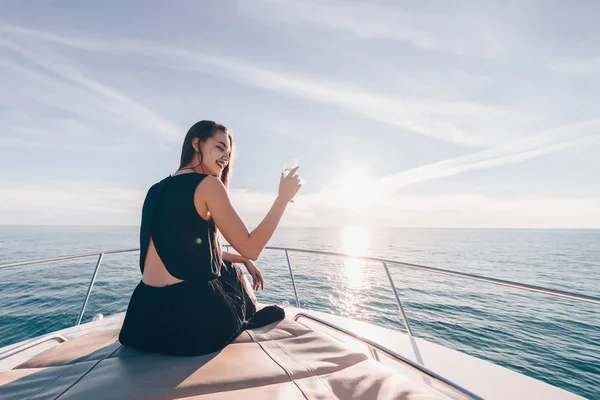 Красивая молодая женщина в черной летней одежде отдыхает на яхте, пьет шампанское и смеется — стоковое фото