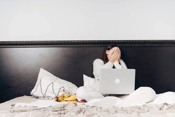 Девушка в белой пижаме сидит на кровати, ест полезные фрукты и смотрит ужасную пленку на ноутбуке, закрывая лицо руками — стоковое фото