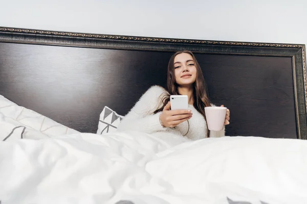 夕方には、ハードな一日の仕事の後休んでいると、スマート フォンを保持ベッドで横になっているパジャマで美しい少女 — ストック写真