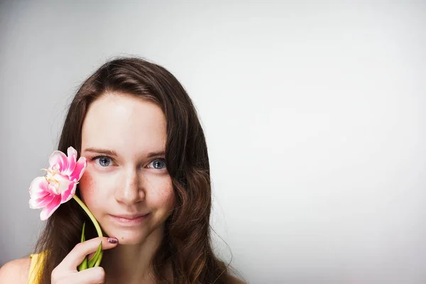 Bela menina de olhos azuis segurando uma flor rosa perfumada perto do rosto e olhando para a câmera — Fotografia de Stock