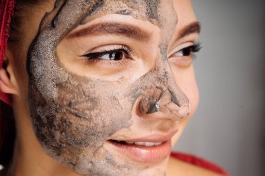 kafasındaki bir havlu ile gülümseyen genç bir kızın yüzüne karşı yararlı kil maskesi uygulanmış, güzel olmak istiyor