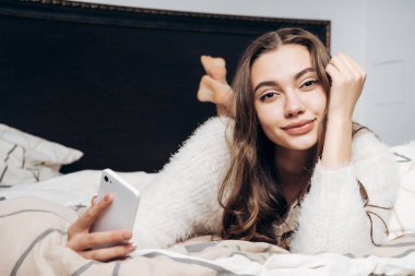 çekici güzel kız dinlenme bir yatağa yatıyor, bir akıllı telefon tutar ve kameraya doğru görünüyor