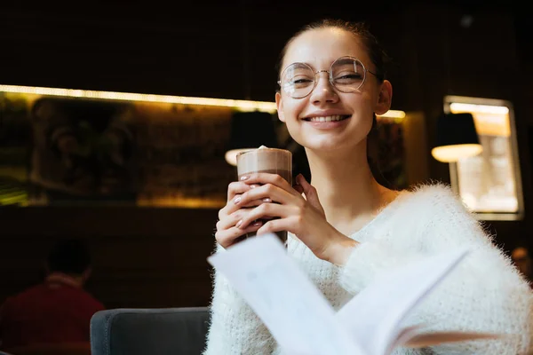 Милая юная студентка в очках отдыхает в кафе после школы и улыбается, пьет капучино — стоковое фото