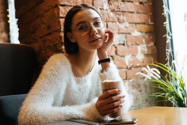 Szemüveg a gyönyörű fiatal nő ül egy kávézóban, és italok egy finom latte, gondolkodik munka — Stock Fotó
