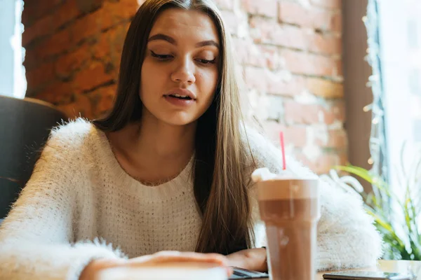 Милая длинноволосая девушка-фрилансер в белом свитере сидит в кафе, работает и пьет ароматный капучино — стоковое фото