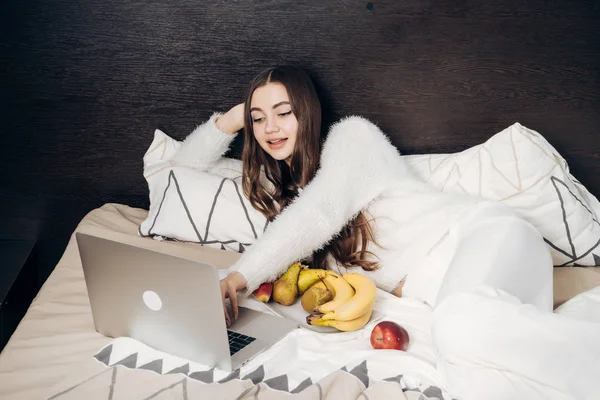 Mooie langharige meisje in pyjama ligt in bed laat op de avond, kijken naar een interessante Tv-show en het eten van fruit — Stockfoto