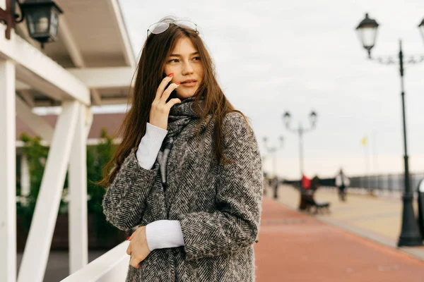 Jolie fille brune urbaine en manteau gris à la mode marchant, parlant au téléphone et souriant — Photo