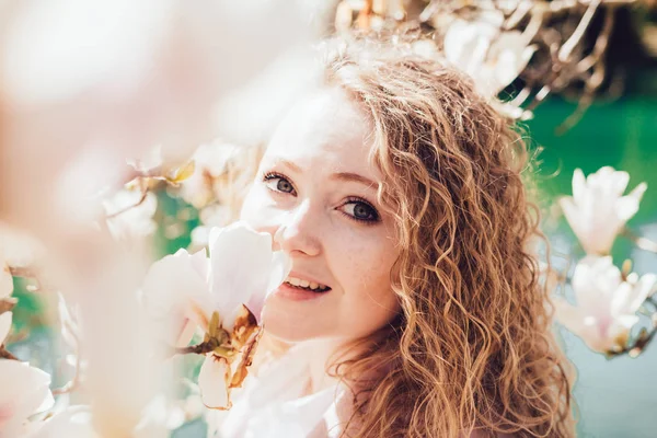 Feliz bela mulher encaracolado posando ao lado de magnólia florescente no parque e sorrindo — Fotografia de Stock