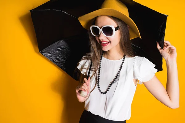 Confiante jovem modelo em chapéu de moda e óculos de sol guarda guarda-chuva e posando no fundo laranja — Fotografia de Stock