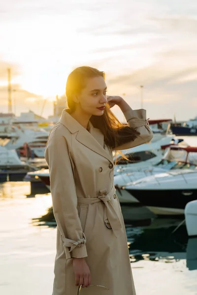 Une jeune femme luxueuse aux cheveux longs, vêtue d'un manteau beige à la mode, se tient dans le port le soir et attend son navire — Photo