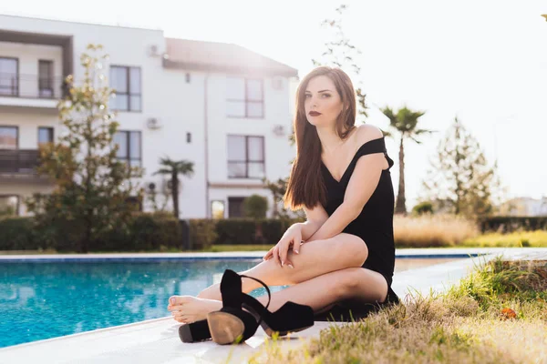 Hermosa chica sexy en un vestido largo negro está descansando junto a la piscina azul en el sol y sonriendo — Foto de Stock