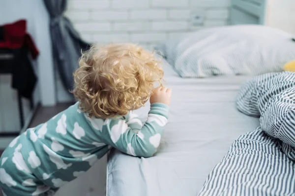 small curly blonde baby boy in blue pajamas stands by the bed, woke up