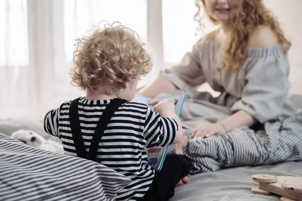 Kręcone mały chłopiec dziecko siedząc na łóżku i grając obok Mama zabawy — Zdjęcie stockowe