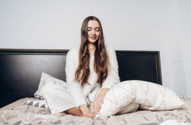 güzel uzun saçlı Genç kız beyaz pijamalı sabah erkenden uyanıp, yatak ve gülümser oturur