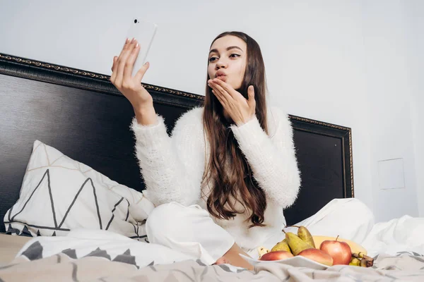 Милая длинноволосая девушка в белой пижаме сидит на кровати, делает селфи и посылает воздушный поцелуй — стоковое фото