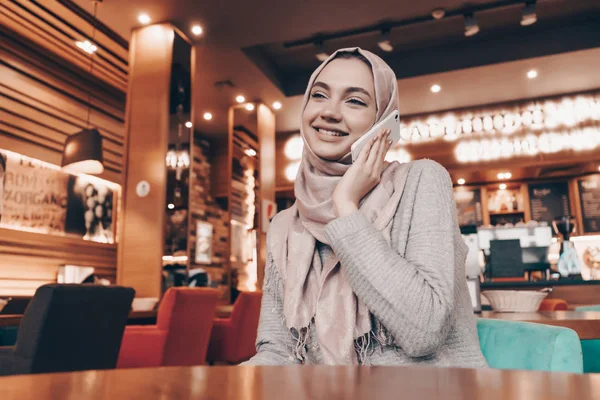 Tersenyum gadis muslim cantik dengan kerudung di kepalanya berbicara di telepon, duduk di kafe — Stok Foto