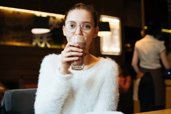 Молодая девушка студентка в очках и белая куртка пьет вкусный сладкий кофе в кафе — стоковое фото