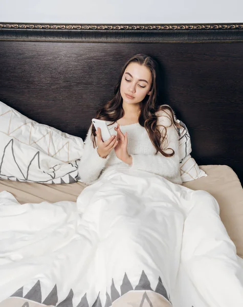 Joven somnolienta en pijama blanco se encuentra en la cama, pone un despertador en su teléfono inteligente — Foto de Stock