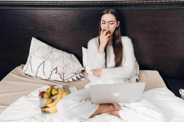 Langharige meisje in pyjama zitten in bed in de avond, rusten en kijken naar een interessante Tv-show op haar laptop, eten van een appel — Stockfoto