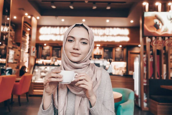 Una mujer musulmana linda con un pañuelo en la cabeza bebe un café caliente fragante en un restaurante, mira a la cámara y sonríe — Foto de Stock