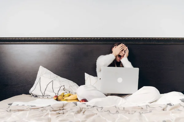 Dziewczyna w białej piżamy siedzi w łóżku, oglądanie scary movie na swojego laptopa, zasłaniając twarz w dłoniach — Zdjęcie stockowe