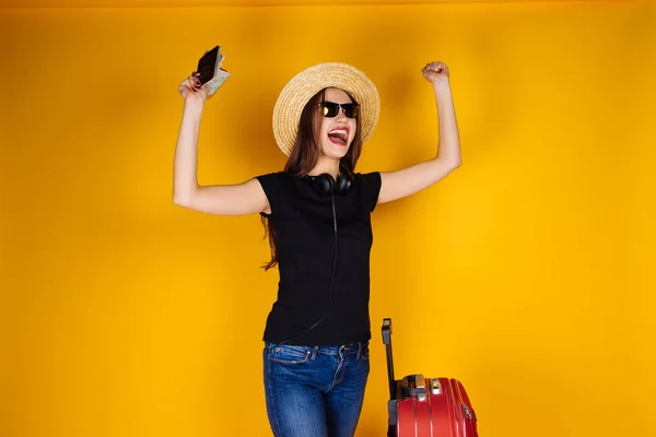 Счастливая молодая девушка в шляпе и очках отправляется в отпуск с большим красным чемоданом, на самолете — стоковое фото