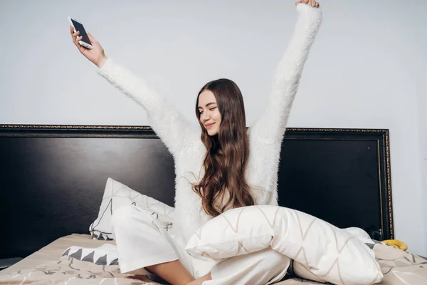 Mooie langharige meisje in pyjama zit op het bed en strekt zich uit, vroeg in de ochtend wakker — Stockfoto