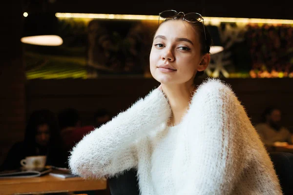 Красива молода дівчина-фрілансер в білому светрі відпочиває в кафе від роботи, дивлячись на камеру — стокове фото