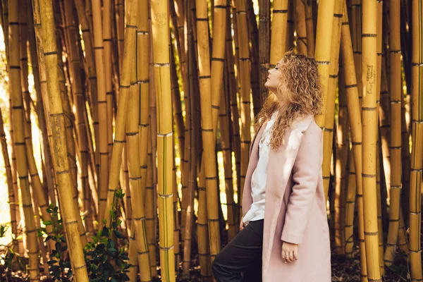 Stylish young woman in trendy pink coat posing in park next to bamboo trees — Stock Photo, Image