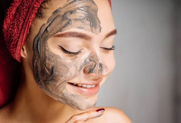 başında kırmızı bir havlu ile çok hoş bir gülümseyen kız bir kil maskesi için yarım yüz uygulanan.