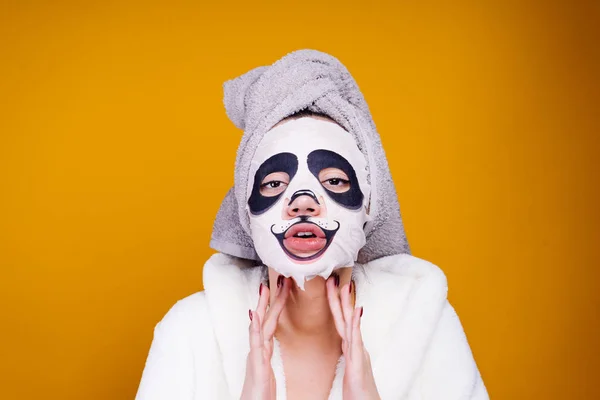 Una joven divertida con una bata blanca y con una toalla en la cabeza cuida de sí misma, en su cara una máscara hidratante con una cara de panda — Foto de Stock