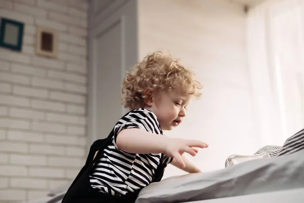 En liten ljushårig pojke i söta kläder vill klättra upp på sängen — Stockfoto