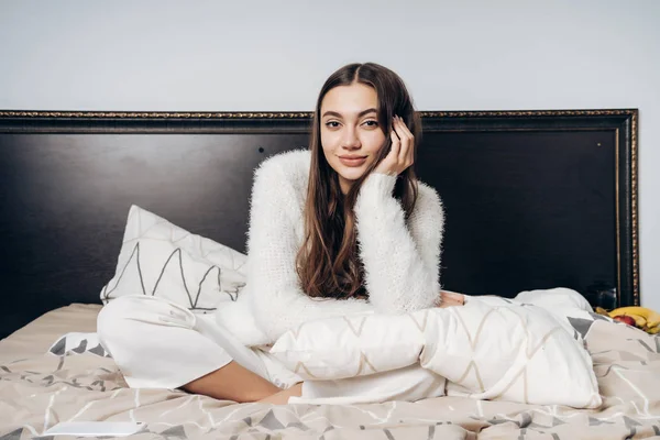 Roztomilý ospalá dívka v pyžamu, sedí na posteli brzy ráno, s úsměvem a při pohledu na fotoaparát — Stock fotografie