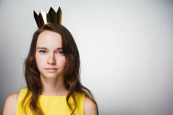 Seria chica de ojos azules jóvenes en un vestido amarillo mira a la cámara, en su cabeza una corona de oro — Foto de Stock