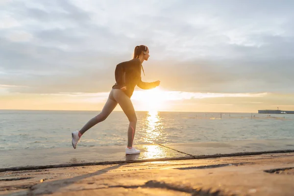Självsäker snabb tjej jogging vid havet i solnedgången, lyssna på musik i hörlurar — Stockfoto
