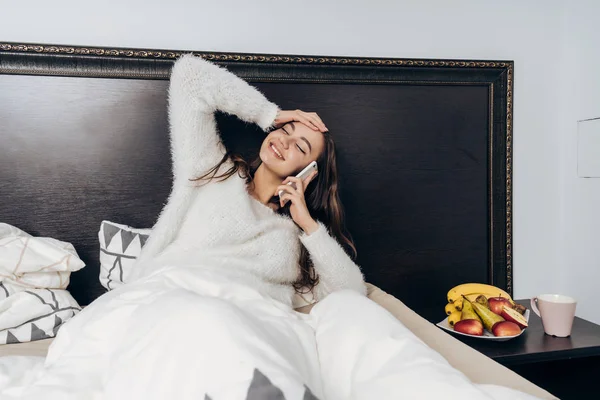 Fiatal lány nevetve fehér pizsamában fekszik az ágyban reggel, beszél a telefonon, jó hangulat — Stock Fotó