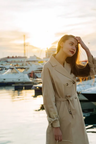 Atrakcyjny dziewczynka długowłosy w stylowy płaszcz beżowy pozowanie w seaport o zachodzie słońca — Zdjęcie stockowe
