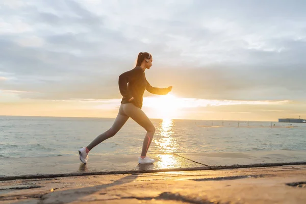 Chica deportiva confiada trotando junto al mar al atardecer, escuchando música en los auriculares — Foto de Stock