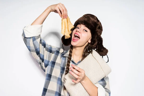 Una divertente ragazza russa con le trecce, in un cappello di pelliccia mangiare frittelle calde deliziose e celebrare Shrovetide — Foto Stock